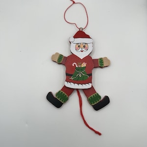 May include: A wooden Santa Claus ornament with a red suit, white beard, and green sack graphic on the chest. The figure has articulated limbs, green cuffs, black boots, and a red string loop at the top for hanging, with a red pull string at the bottom.