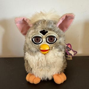 May include: A plush toy featuring long, shaggy gray and white fur, large brown eyes with black lashes, and prominent pink-lined ears. It has a bright yellow beak, orange feet, and a small purple tag attached to the side with a graphic illustration.