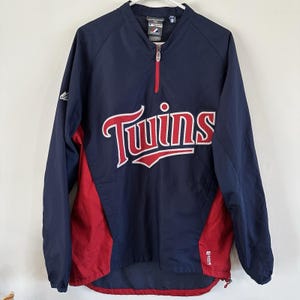 May include: A navy blue and red long-sleeve pullover jacket featuring a quarter-zip neckline and the word Twins printed in red with white trim across the chest. The garment includes a Majestic brand logo on the right sleeve and a small tag at the neck.