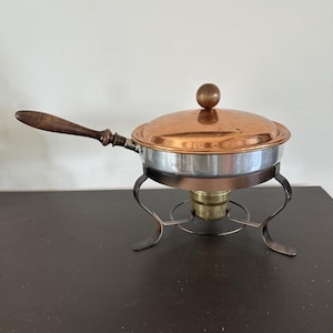 Peut inclure: Un plat à réchaud en métal cuivré et argenté avec un couvercle rond muni d'un bouton en bois. La poêle possède un long manche en bois foncé et repose sur un support en métal sombre avec une coupelle brûleur en laiton au centre.