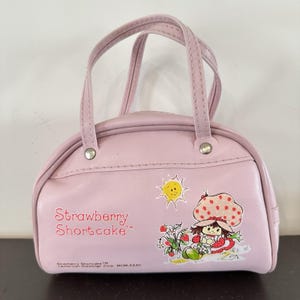 May include: A light pink handbag featuring a graphic of the character Strawberry Shortcake, a yellow sun, and strawberry plants. The text reads Strawberry Shortcake and includes the copyright information American Greetings Corp. MCMLXXXII.