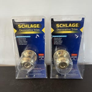 May include: Two Schlage Decorative Trim door knobs in brass finish, each encased in clear plastic retail packaging. The blue and yellow backing cards feature the Schlage logo, a list of suggested applications, and a full lifetime mechanical warranty.
