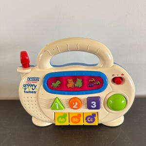 May include: A cream-colored VTech Little Smart Groovy Tunes toy with a handle, featuring a red antenna, a blue speaker grille, and a central display showing a green insect, two bears, and two cars. It includes buttons labeled 1, 2, 3, Aa, Bb, and Cc.