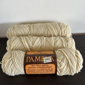 Op de afbeelding: Drie strengen gebroken wit Lion Brand Pamela breigaren horizontaal gestapeld. Het middelste label vermeldt 100% maagdelijk acryl, kamgaren gewicht, 4-draads, 3,5 oz, 99,2 gram nettogewicht, machinewasbaar en geschikt voor de droger.