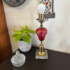 May include: A table lamp featuring a square marble base, a brass bell-shaped stand, and a red glass body with etched grape designs. The lamp has a brass socket and a white light bulb. Beside it sits a clear, round, lidded glass trinket dish.