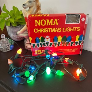 Może przedstawiać: Pudełko lampek choinkowych NOMA Christmas Lights zawierające 15 żarówek do użytku wewnętrznego, z czerwonym tłem i żółtym napisem. Przed pudełkiem na ciemnej powierzchni rozłożony jest sznur wielokolorowych żarówek w kolorach czerwonym, niebieskim, zielonym, pomarańczowym i białym.