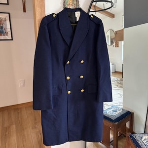 Puede incluir: Un abrigo cruzado de color azul marino con botones dorados, charreteras en los hombros y cuello de solapa. El abrigo presenta una silueta estructurada con dos bolsillos con solapa en la parte delantera y forro negro visible en el cuello.