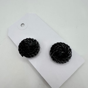 May include: A pair of circular stud earrings featuring a dense, layered arrangement of small, shiny black sequins. The earrings are attached to a white rectangular display card with a circular hole at the top for hanging, providing a clear view of the textured, reflective surface of the black circular components.