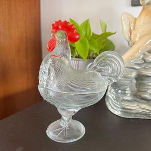 Puede incluir: Un plato con tapa de vidrio prensado transparente con forma de gallo, que presenta un patrón de plumas texturizado en el cuerpo y una cresta y barbillas de vidrio rojo vibrante en la cabeza. El gallo se asienta sobre una base de pedestal.
