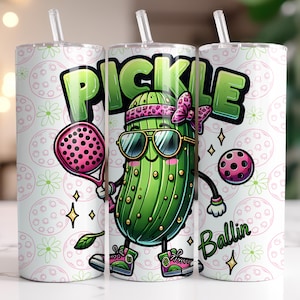 May include: A white tumbler with a green pickle cartoon holding a pink paddle and a pink ball. The pickle is wearing sunglasses and a pink bow. The tumbler has a white background with a repeating pattern of green leaves and pink circles. The text "Pickle Ballin'" is printed on the tumbler.