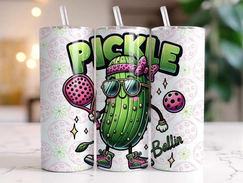 May include: A white tumbler with a green pickle cartoon holding a pink paddle and a pink ball. The pickle is wearing sunglasses and a pink bow. The tumbler has a white background with a repeating pattern of green leaves and pink circles. The text "Pickle Ballin'" is printed on the tumbler.