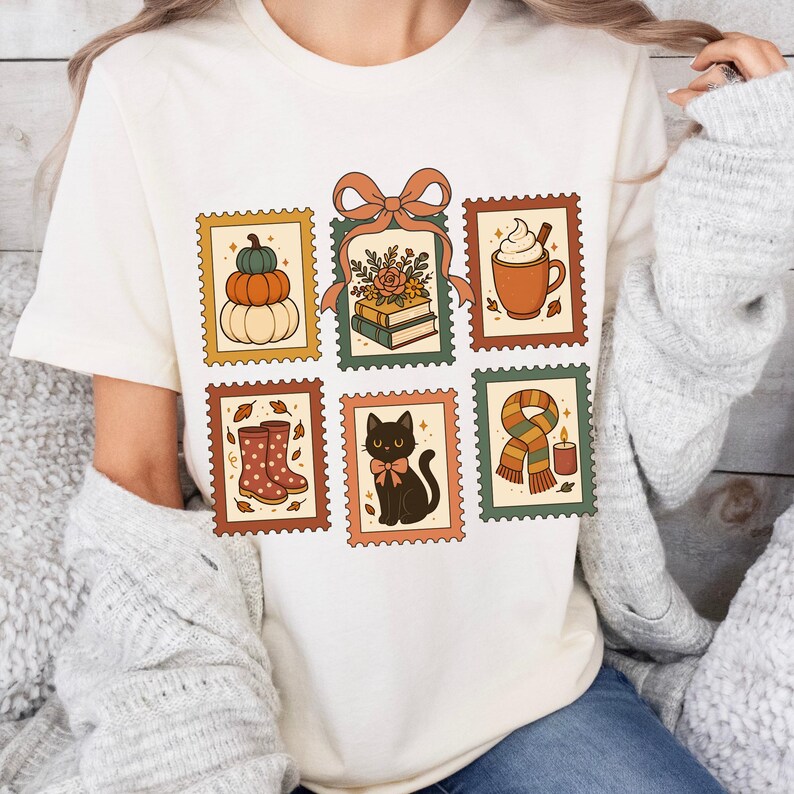 May include: Cream-colored t-shirt featuring six postage stamp-style illustrations with fall themes. Designs include pumpkins, books with flowers, a coffee mug, rain boots, a black cat, and a scarf with a candle.