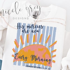 May include: White sweatshirt with a graphic design featuring the text "His mercies are new every morning" and "Lamentations 3:22-23". The design includes a sunrise with blue, orange, and pink colors. A gold necklace is draped across the sweatshirt.
