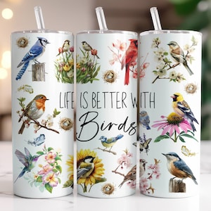 May include: Three white stainless steel tumblers with lids and straws. Each tumbler features a different colourful bird design with flowers and leaves. The middle tumbler has the text "Life is better with birds".