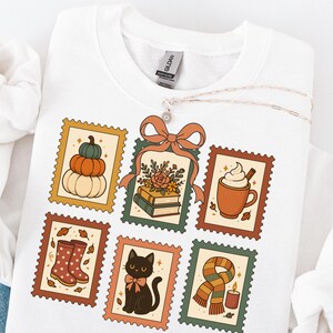 May include: White sweatshirt featuring six postage stamp designs with fall themes. Designs include pumpkins, books, a coffee mug, rain boots, a black cat, and a scarf with a candle. A silver necklace with a flower pendant is draped over the sweatshirt.