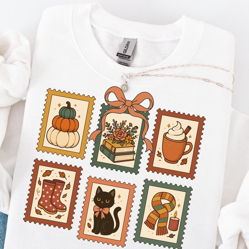 May include: White sweatshirt featuring six postage stamp designs with fall themes. Designs include pumpkins, books, a coffee mug, rain boots, a black cat, and a scarf with a candle. A silver necklace with a flower pendant is draped over the sweatshirt.