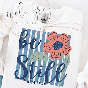 May include: White sweatshirt with a floral design and the words "Be Still" in navy blue script. The design includes a coral flower, green leaves, and blue and white striped background. The text "Psalm 46:10" is also included.