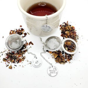 May include: Two silver tea infusers with chains and round metal tags. The tags have engraved sayings: "I Like Big Mugs & I Cannot Lie", "No Teabagging", and "Best Tea Friend". The infusers are on a white surface with loose leaf tea scattered around them.