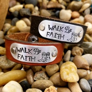 May include: Two brown leather bracelets with silver metal plates that say "Walk By Faith" and have a small boot design. The bracelets are on a bed of small brown rocks.