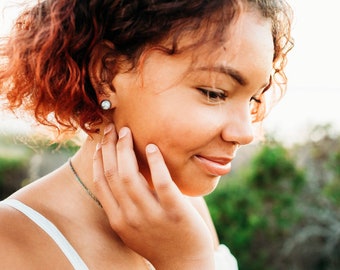 May include: A young woman with curly red hair is wearing a white tank top and a silver necklace. She is touching her cheek with her hand and looking down. She is wearing a small, round earring in her left ear.