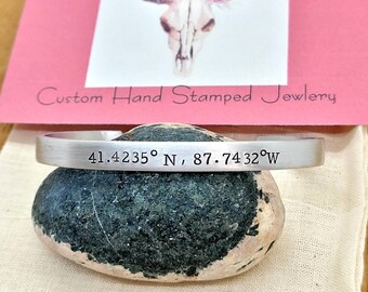 May include: Silver bracelet with the coordinates 41.4235° N, 87.7432° W engraved on it. The bracelet is resting on a gray rock.
