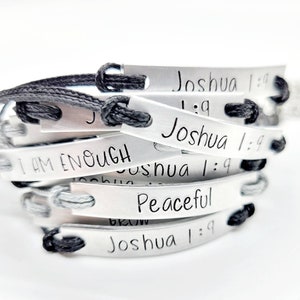 Peut inclure: Une pile de bracelets en argent avec des cordons noirs. Les bracelets ont différents messages inspirants gravés dessus, dont "Je suis assez" et "Paisible". Certains bracelets incluent également le texte "Josué 1:9".