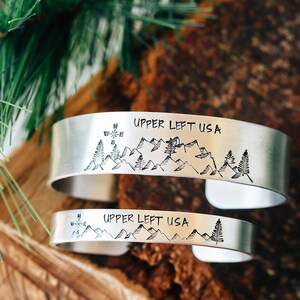 May include: Two silver bracelets with a mountain range and compass design. The bracelets are engraved with the text "Upper Left USA".