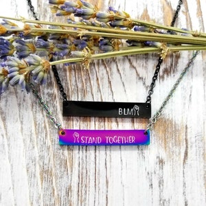 May include: Two silver necklaces with black and rainbow bar pendants. The black pendant has the text "BLM" and a raised fist symbol. The rainbow pendant has the text "I STAND TOGETHER" and a raised fist symbol.