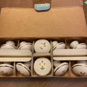 Könnte beinhalten: Ein Set aus weißen Vintage-Keramik-Schrankknöpfen mit goldenen Akzenten und einem Fleur-de-Lis-Design, angeordnet in einer Pappschachtel. Die Schachtel hat ein blaues Etikett mit der Aufschrift "Towne".