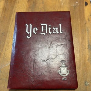 May include: A maroon, textured hardcover book titled Ye Dial in white gothic lettering. The cover features a silver emblem with the year 1766 above it and the year 1961 below it. The book has a leather-like finish with visible surface creases.