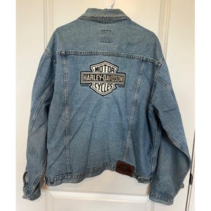 Könnte beinhalten: Hellblaue Jeansjacke mit dem Harley-Davidson Cycles-Logo auf dem Rücken. Die Jacke hat ein klassisches Design mit Knopfleiste, Kragen und Taschen. Ein brauner Lederaufnäher befindet sich auf der unteren rechten Seite.