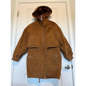 May include: A brown corduroy jacket with a hood, zipper closure, and two front pockets. The jacket has a drawstring at the waist and cuffs. The jacket is hanging on a white door.