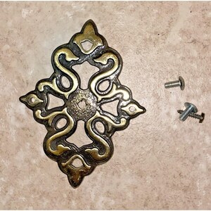 May include: Ornate, antique-style brass cabinet pull with a swirling design. The decorative hardware features a central square and is accompanied by two screws for installation. Ideal for furniture restoration.