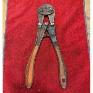 Op de afbeelding: Een vintage metalen betonschaar met verweerde donkergrijze stalen bekken en handgrepen met versleten oranje verf. Het gereedschap is voorzien van in reliëf aangebrachte tekst op de handgreep met de tekst The National Telephone Supply Co. Cleveland Ohio.