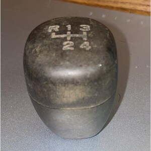 May include: A black manual transmission gear shift knob with a rounded rectangular shape. The top surface features an engraved gear pattern showing R, 1, 3, 2, and 4 positions. The material appears to be a matte, textured plastic or rubber finish.