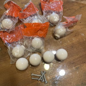 May include: A collection of round, white ceramic cabinet knobs, some individually sealed in clear plastic bags with orange labels, alongside several loose metal mounting screws. The knobs have a smooth, spherical shape and a glossy finish.