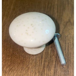 Puede incluir: Pomo de armario color crema con un patrón moteado. El pomo redondo está unido a un tornillo plateado. El pomo está sobre una superficie de madera.