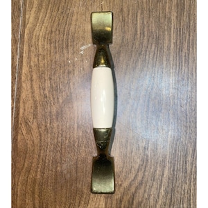 Könnte beinhalten: Ein Schrankgriff mit rechteckigem Messingende, einem weißen ovalen Mittelteil und Messingakzenten. Die Hardware ist für Möbel konzipiert und hat eine Vintage-Ästhetik.