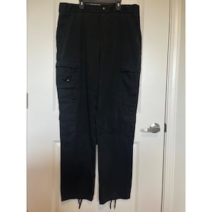 Könnte beinhalten: Schwarze Cargohose mit mehreren Taschen an Beinen und Seiten. Die Hose hat ein gerades Bein-Design und einen Kordelzug am unteren Saum. Die Hose hängt an einem Kleiderbügel vor einer weißen Tür.