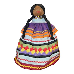 May include: Handmade doll with a brown face, black braided hair, and a beaded necklace. The doll wears a colourful dress with a purple top, orange trim, and a skirt with geometric patterns in red, yellow, and purple.