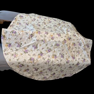 May include: Cream-colored floral patterned fabric with purple and yellow flowers and ribbon accents. The fabric is draped over an unseen object, creating a soft, textured appearance. The floral design is evenly distributed across the surface.