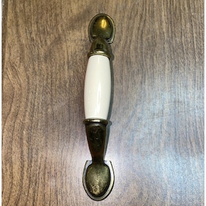 May include: A cabinet pull handle featuring a central, smooth, off-white ceramic cylinder flanked by two aged brass-toned metal end pieces. The metal components have a weathered, dark gold finish with rounded, spade-shaped mounting plates.