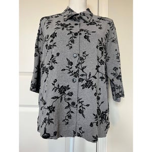 May include: A gray button-down shirt featuring a black floral pattern. The shirt has a classic collar, long sleeves, and a front button closure. The fabric displays a repeating design of delicate flowers and vines across the entire garment.