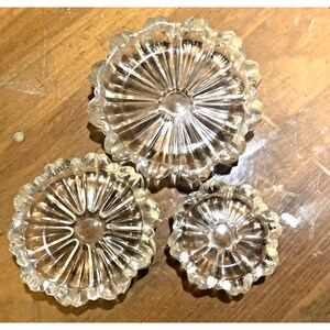 May include: Three clear glass dishes of varying sizes, each featuring a circular center and a radiating sunburst pattern of ridges extending to a scalloped, petal-like edge. The glass is transparent and reflective, showing a distinct radial design.