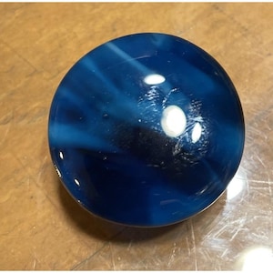 May include: A blue glass marble with white and dark blue swirls. The marble has a glossy finish and a few small air bubbles. The marble is a sphere and is sitting on a wooden surface.