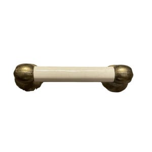 May include: A cabinet pull with a cream-coloured central section and brass-coloured ends. The pull is cylindrical with rounded ends, suggesting a vintage or antique style. The brass ends have a textured, possibly aged, appearance.