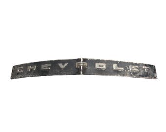 May include: A vintage black metal Chevrolet emblem badge with raised silver-toned lettering. The rectangular piece features a slight center crease and weathered finish, displaying the word CHEVROLET in a bold, block font across the front surface.