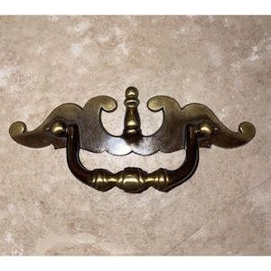 Puede incluir: Tirador de cajón de latón antiguo con un diseño decorativo. El tirador presenta un asa curva y detalles ornamentados, con un asa marrón oscuro y detalles en color latón. El herraje está diseñado para la restauración de muebles o uso decorativo.