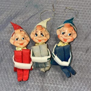 May include: Three vintage Christmas elf ornaments. Each elf has a smiling face, rosy cheeks, and a small hat. One elf wears a red hat and carries a red gift. The other two elves wear hats in white and blue, and carry gray and blue gifts.