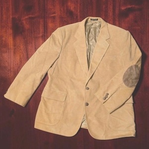 Pode incluir: Um blazer de veludo cotelê bege com lapela entalhada, dois botões frontais, dois bolsos com aba e cotoveleiras marrons contrastantes nas mangas. O casaco inclui um forro interno liso e três pequenos botões em cada punho da manga.